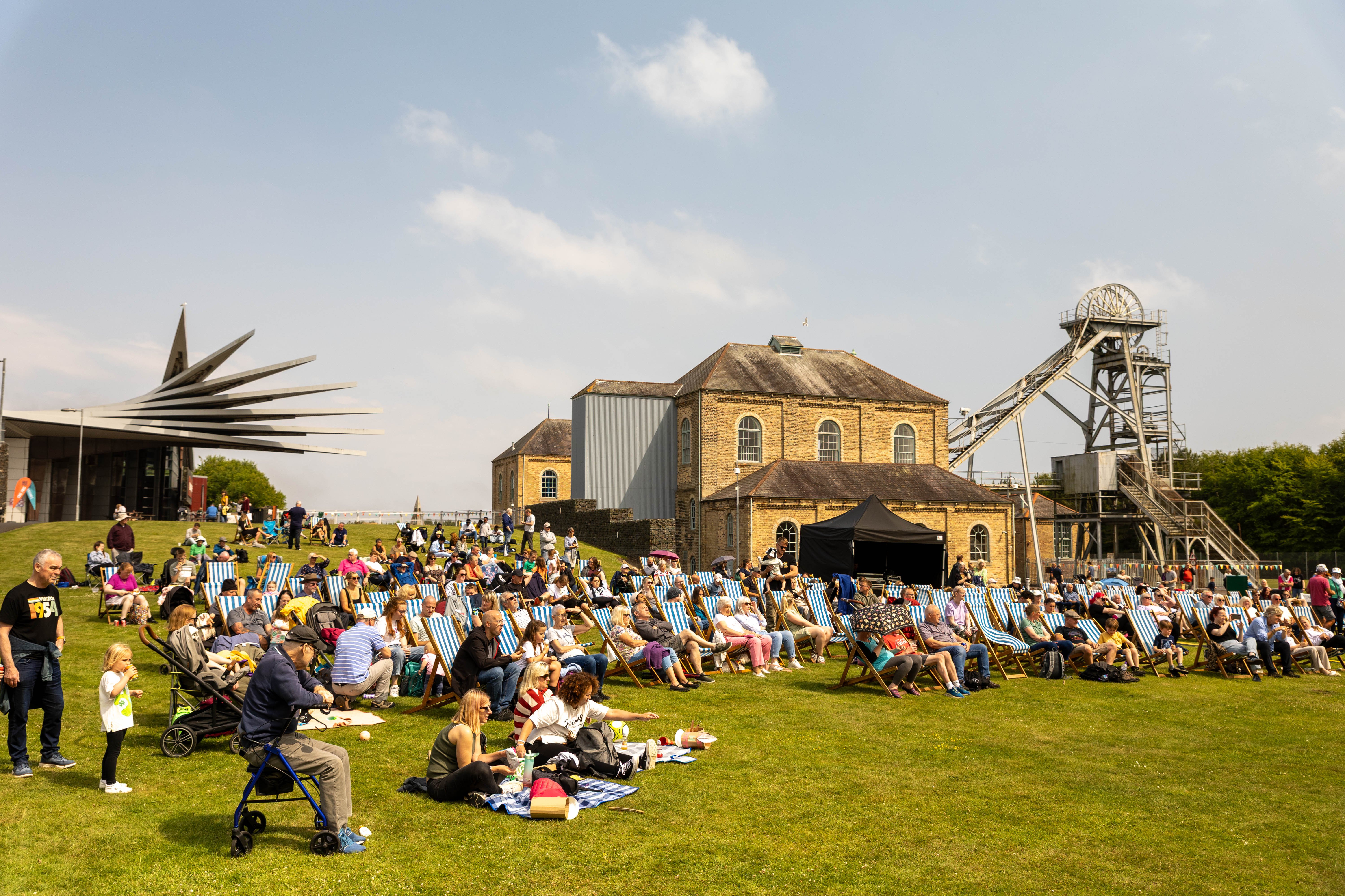 An exterior view of Woodhorn Museum where a crowd of people are sat outside on picnic chairs for Northumberland Miners' Picnic Day