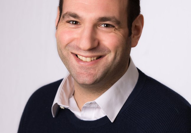 smiling brown haired man in dark jumper & white shirt 