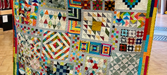 A multicoloured fabric quilt held between two hands. The quilt features a variety of shapes and fabrics.