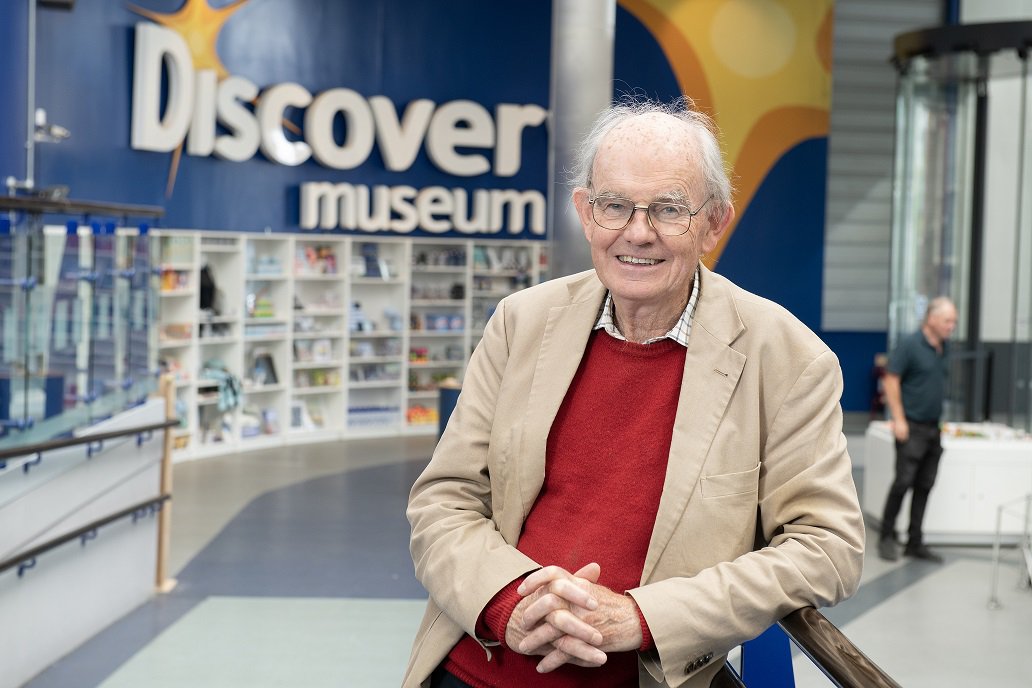 A man smiling and leaning on a railing with a large sign behind him reading Discovery Museum A man smiling and leaning on a railing with a large sign behind him reading Discovery Museum