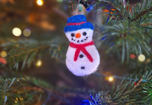 A photograph of a needle felted snowman Christmas decoration hanging on a Christmas tree.