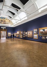 A photograph showing a gallery space with dark blue walls and paintings of all different sizes and frames hung on the walls. 