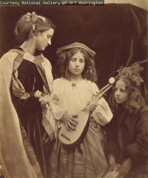 A sepia photograph of three children standing or seated in front of a dark background. They are wearing historical costumes. The child in the centre is holding a lute or similar instrument.